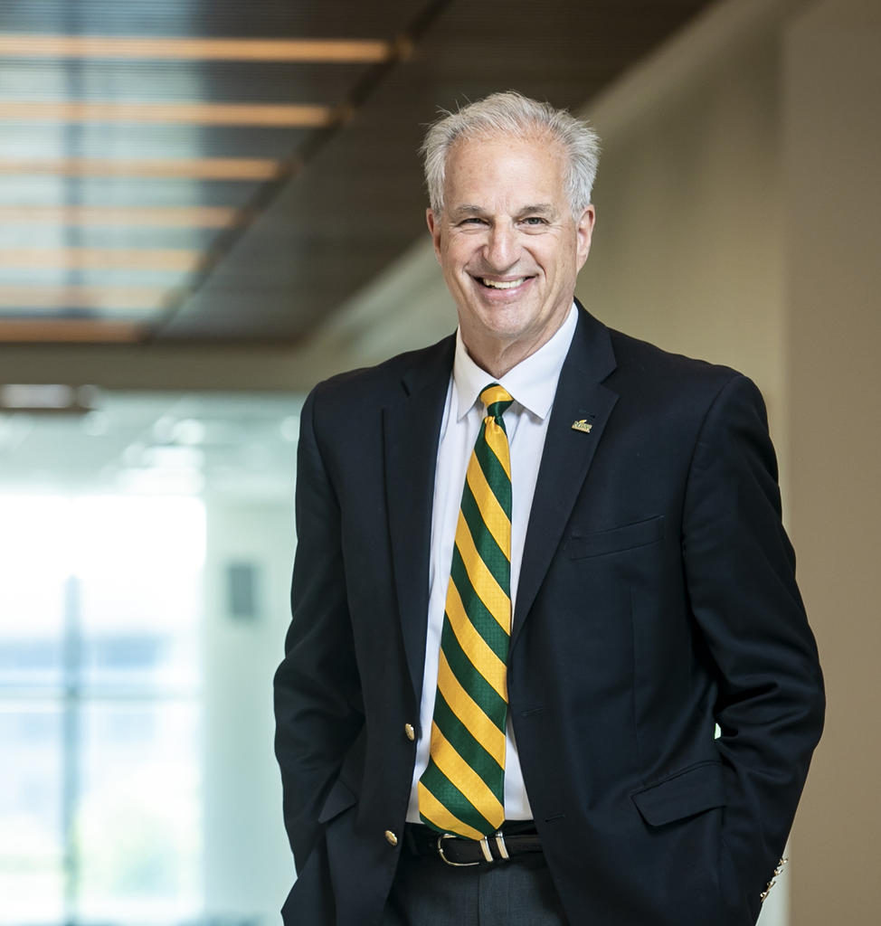 Smiling man with gray hair wearing dark colored suit with green and gold stripe tie over a white shirt.