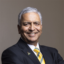 Gray-haired, smiling man wearing a black suit with striped yellow tie.