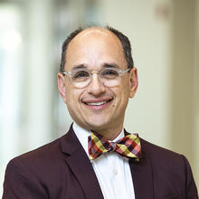 Smiling man with brown hair wearing glasses and a burgundy jacket with plaid bow tie.