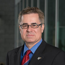 Short-haired man wearing black glasses and suit with red, striped tie.