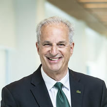 Man with gray hair wearing a black suit, white shirt, and dark green tie.